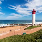 Paved Green vegetation and patterned walkway with metal barrier against red and white lighthouse against blue cloudy coastal seascape at Umhlanga, Durban, South Africa