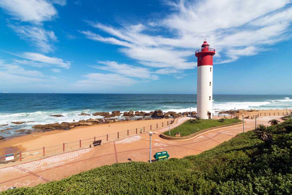 Paved Green vegetation and patterned walkway with metal barrier against red and white lighthouse against blue cloudy coastal seascape at Umhlanga, Durban, South Africa